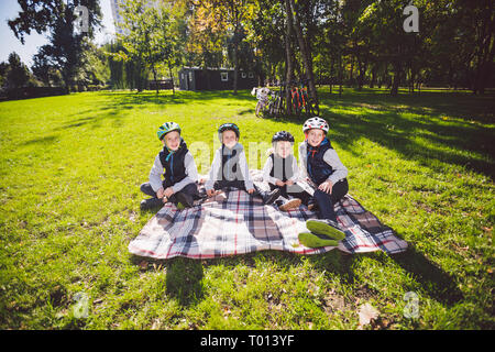 Große familie Kaukasischen Kindern. Drei Brüder und Schwester sitzen auf Decke außerhalb des Parks. Grüne wiese gras Hintergrund von Fahrrädern. Lustig Stockfoto
