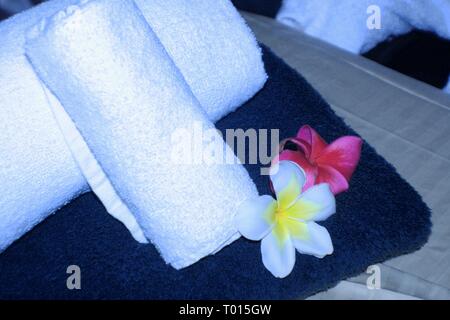 Nahaufnahme eines Massagetisches mit weißen Rollhandtüchern, blauem Handtuch und dekoriert mit roten und weißen Plumeria-Blumen in einem Spa Stockfoto