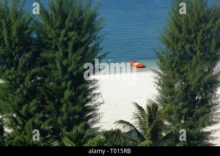 Ein rot-gelber Jetski bildet einen schönen Kontrast, der am blauen Wasser und am weißen Sandstrand, eingerahmt von grünen Bäumen, parkt Stockfoto