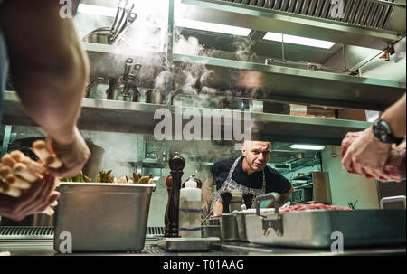 Ist alles ok Surious Chef ist auf der Suche, wie man seine Assistenten sind Kochen Fleisch in einem Restaurant Küche. Kulinarische Schule Stockfoto