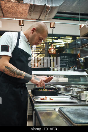 Finishing ein Gericht. Seitenansicht des jungen männlichen Chef mit mehreren Tattoos auf seinem Arme garnieren italienische Pasta in einem Restaurant Küche. Stockfoto