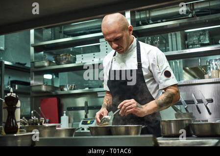 Fast fertig. Portrait von gutaussehenden Koch in schwarze Schürze und mit mehreren Tattoos auf seinem Arme Kochen einer Mahlzeit in seinem Restaurant Küche. Kochen Stockfoto