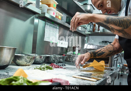 Die pasta-Prozess. Nahaufnahme Foto von konzentriertem Küchenchef mit schwarzen Tattoos auf seinem Arme gießen Mehl am Küchentisch, während Sie Pasta. Kochen Stockfoto