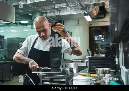 Hausgemachte Pasta. Portrait von konzentrierten jungen Koch rollen einer schwarzen Teig durch pasta Maschine im Restaurant Küche. Essen Konzept Stockfoto