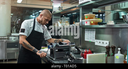 Bei der Arbeit konzentriert. Ernst und gutaussehenden Koch mit schwarzen Tattoos auf seine Hände Rollen einer schwarzen Teig durch pasta Maschine. Italienische Pasta Stockfoto