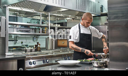 Fast fertig. Stattliche professioneller Koch mit Tattoos auf seinem Arme mischen einen Salat in einer Metallschüssel, während in der Küche stehen. Kochen Stockfoto