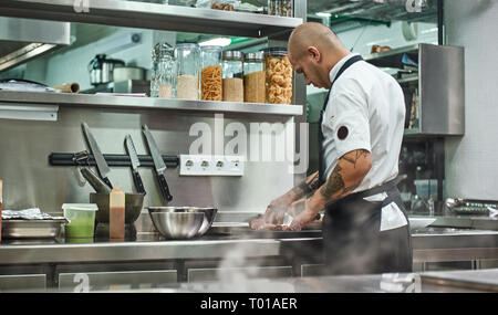 Arbeitsprozess. Rückansicht des männlichen Chef in Schürze und mit Tattoos auf seinem Arme Schneiden von Fleisch, während in einem Restaurant in der Küche stehen. Kulinarische Konzept Stockfoto