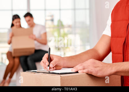 Nahaufnahme einer Lieferung Mann in Rot Post gleichmässige Füllung in Dokument, halten es auf einem Karton. Glückliches Paar auspacken ihre Paket im Hintergrund. Beschäftigte Arbeitnehmer, hohe Qualität. Im Innenbereich. Hellen Innenraum. Stockfoto
