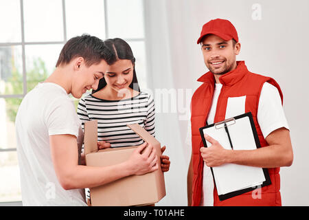 Fröhliche Postbote das Tragen der roten Post einheitlich ist die Bereitstellung von Parzellen zu zufriedenen Kunden. Er bringt Kartons an seinen Besitzer. Er hat seinen Job sehr gut gemacht. Dunkelhaarige Frau und jugendlich Öffnen Paket, sie freuen sich, Sie zu empfangen. Freundliche Mitarbeiter, hohe Qualität. Im Innenbereich. Hellen Innenraum. Stockfoto