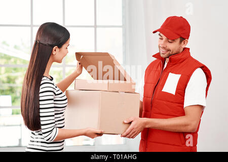Fröhliche Postbote das Tragen der roten Post einheitlich ist die Bereitstellung von Parzellen zu einem zufriedenen Kunden. Er bringt Kartons an seinen Besitzer. Dunkelhaarige Frau öffnet Paket, Sie ist froh, Sie zu empfangen. Freundliche Mitarbeiter, hohe Qualität. Im Innenbereich. Hellen Innenraum. Stockfoto