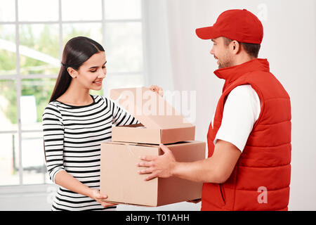Fröhliche Postbote das Tragen der roten Post einheitlich ist die Bereitstellung von Parzellen zu einem zufriedenen Kunden. Er bringt Kartons an seinen Besitzer. Dunkelhaarige Frau öffnet Paket, Sie ist froh, Sie zu empfangen. Freundliche Mitarbeiter, hohe Qualität. Im Innenbereich. Hellen Innenraum. Stockfoto