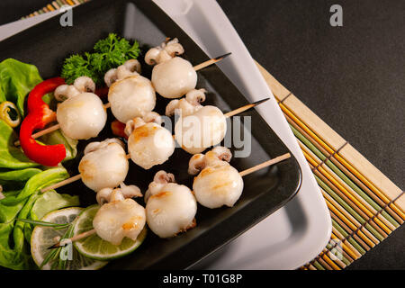 Baby Tintenfisch Grill auf einer hölzernen Spieß in einer schwarzen Platte Stockfoto