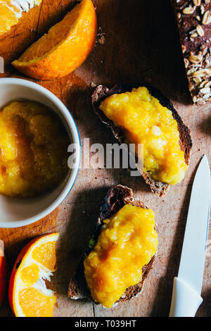 Hausgemachte Orange Marmelade auf einem natürlichen Rohstoff Weizen Brot und Orangenscheiben über eine hölzerne cutboard, gesundes Frühstück, in der Nähe und windom Licht. Stockfoto