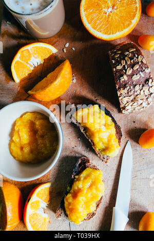 Hausgemachte Orange Marmelade auf einem natürlichen Rohstoff Weizen Brot und Orangenscheiben über eine hölzerne cutboard, gesundes Frühstück, in der Nähe und windom Licht. Stockfoto