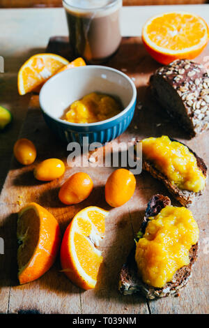 Hausgemachte Orange Marmelade auf einem natürlichen Rohstoff Weizen Brot und Orangenscheiben über eine hölzerne cutboard, gesundes Frühstück, in der Nähe und windom Licht. Stockfoto