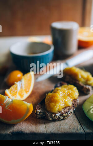 Hausgemachte Orange Marmelade auf einem natürlichen Rohstoff Weizen Brot und Orangenscheiben und eine Tasse Kaffee und Milch über eine hölzerne cutboard, gesundes Frühstück, in der Nähe ein Stockfoto