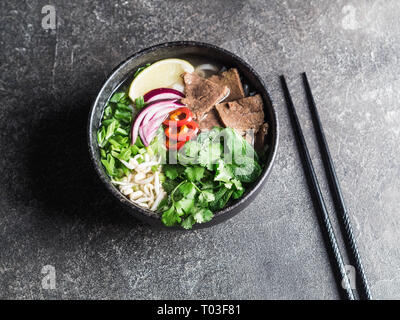 Traditionelle vietnamesische Suppe pho bo von Rindfleisch, Reis Nudeln und Grüns in einem schwarzen Schüssel auf einem grauen Hintergrund. Asiatische Lebensmittel Konzept. Ansicht von oben Stockfoto