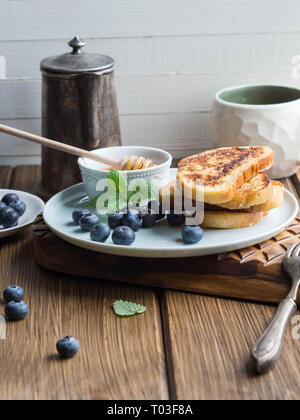 Ein Stapel von hausgemachten French Toast mit Heidelbeeren und Honig auf einem blauen Platte auf einer hölzernen Textur board bestreut. Leckeres Frühstück. Stockfoto