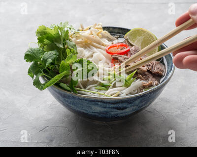 Traditionelle vietnamesische Suppe pho in eine blaue Schale, serviert mit Minze und cilantro, grüne Zwiebeln, Chili, Sojasprossen und Kalk. Hand mit einem Essstäbchen Stockfoto