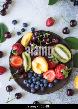 Die verschiedenen Beeren und Früchte auf einer Holzplatte auf grauem Hintergrund. Erdbeeren, Kirschen, Kiwi, Pfirsich, Blaubeeren auf einem Teller. Ansicht von oben Stockfoto