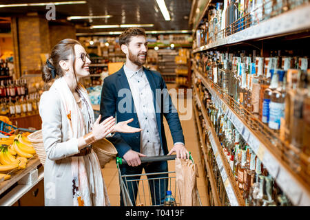 Junger Mann und Frau sie ein starkes Getränk, stehend in der Nähe der Regale mit Alkohol im Supermarkt Stockfoto