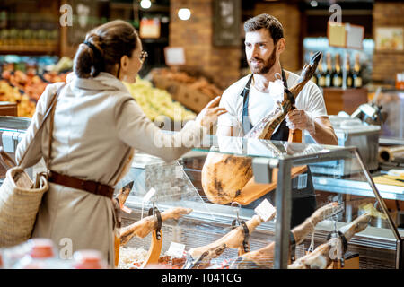 Verkäufer Beratung jamon für eine junge Frau, die Kunden an der Theke stehend mit getrocknetem Fleisch im Supermarkt Stockfoto