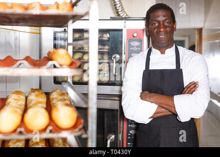 Zuversichtlich Baker in schwarze Schürze mit Waffen in der Nähe von professionellen Brotbackofen in kleine Bäckerei gekreuzt Stockfoto