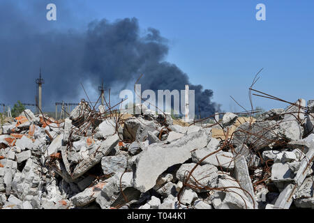 Ein Haufen von betonschutt mit hervorstehenden Armierungseisen auf dem Hintergrund von dichtem schwarzem Rauch in den blauen Himmel. Hintergrund. Das Konzept der Folgen Stockfoto