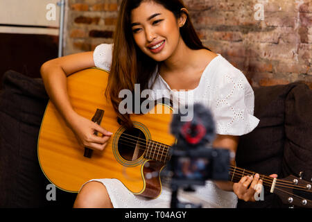 Schöne asiatische Frau lächelnd und spielen Cover Song beim Sitzen auf der Couch vor der Videokamera Stockfoto