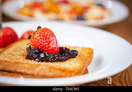 Tischplatte in der Nähe von Brot Toast mit dem Belag der Cranberry Konfitüre und Erdbeere im weißen Platte am Esstisch aus Holz. Stockfoto