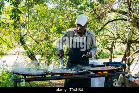 Schwarzer Mann kochen Frühstück auf Outdoor Grill mit Speck und Würstchen zum Frühstück für Safari Gäste, Sabi Sands Game Reserve, Südafrika Stockfoto