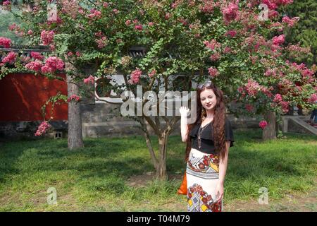 Kaukasische Dame mit langen brünetten Haaren und modische Kleidung vor rosa Blüten auf einem Baum posing Stockfoto