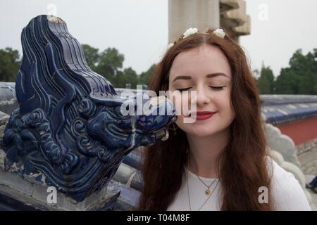 Ruhigen westlichen kaukasischen brunette weibliche Touristen. Ruhige Gesicht, Augen, geschlossen, Blume Krone, sie steht neben einem lächelnden Chinesischen Blue Dragon Carving Stockfoto