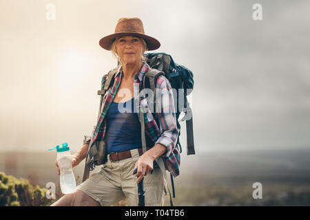 Porträt einer älteren Frau auf einem Wanderweg Trinkwasser auf einem Hügel. Nahaufnahme von einer älteren Frau Wanderer ihre wanderausrüstung stehen auf Stockfoto