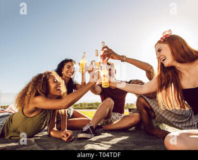 Gruppe von Jugendlichen feiern mit Bier im Freien. Multirassischen Männer und Frauen sitzen zusammen und toasten Biere. Stockfoto
