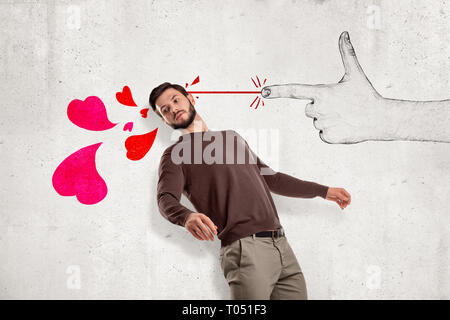 Jungen gutaussehenden Mann in Freizeitkleidung in der Hälfte stehend - Schalten Sie in der Nähe von Mauer und lehnte sich zurück, als ob von Hand gezeichnete finger Gewehr schießen rosa Herzen ihn an. Stockfoto