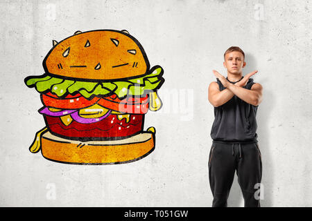 Junge passen Mann in Sleeveless Hoody stehend mit Arme über der Brust an der weißen Wand gekreuzt mit Zeichnung der riesigen Hamburger auf. Stockfoto