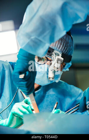 Arzt während der Operation in den Betrieb. Sie trägt eine blaue Maske, bunten Hut und Brille. Sein Assistent ist neben ihm. Close Up. Horizontale. Stockfoto