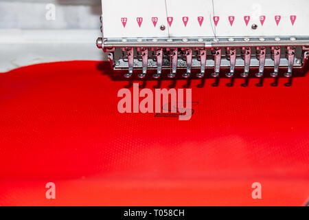 Industrielle weiße Nähmaschine für Stickerei macht schwarzem Logo mit dem Buchstaben S mit schwarzen Faden auf einem roten Hintergrund aus perforiertem Leder Stockfoto