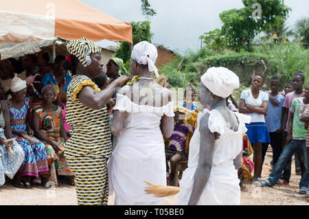 Adzopé, Elfenbeinküste - 31. August 2016: reife Frau in Weiß Halskette Geben zurück zu der Kamera gekleidet, während ihre Schwester im lendenschurz Hals reinigt, Stockfoto
