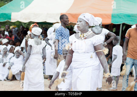 Adzopé, Elfenbeinküste - 31. August 2016: Eine junge Dame in Weiß mit Ketten und eine Glocken Hals gekleidet, ihr Sohn auch in Weiß gekleidet, zu Fuß auf den Tanz Stockfoto