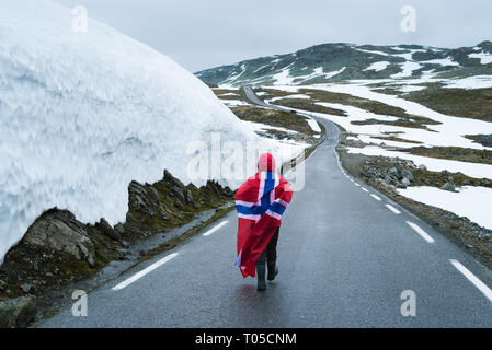 Bjorgavegen - verschneite Straße in Norwegen. Mädchen mit die norwegische Flagge auf einem Berg Straße in der Nähe einer Wand aus Schnee. Schwere nördlichen Landschaft Stockfoto