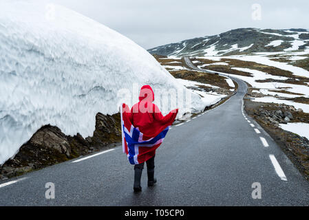 Bjorgavegen - verschneite Straße in Norwegen. Mädchen mit die norwegische Flagge auf einem Berg Straße in der Nähe einer Wand aus Schnee. Touristische Route führt von Aurla Aurlandsfjellet Stockfoto