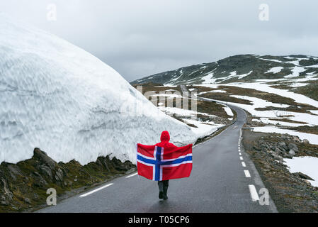 Bjorgavegen - verschneite Straße in Norwegen. Mädchen mit die norwegische Flagge auf einem Berg Straße in der Nähe einer Wand aus Schnee. Touristische Route führt von Aurla Aurlandsfjellet Stockfoto