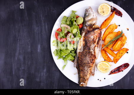 Gebratener Seebarsch mit Zitrone, Kartoffeln, Salat und Gewürzen auf schwarzen Holztisch Stockfoto