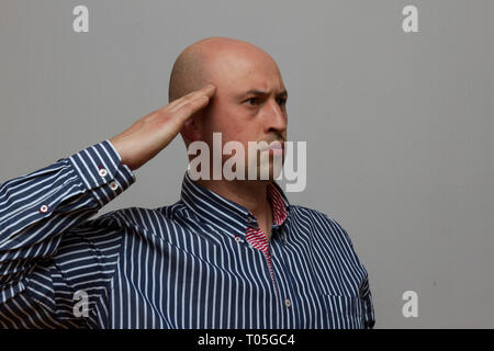 Junge glatzköpfige Mann salutierte auf grauem Hintergrund Stockfoto