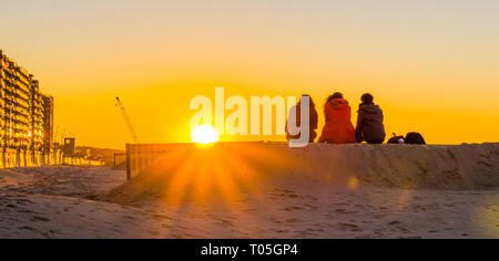 Drei Jugendliche auf einem Sandhügel zusammen sitzen, zuschauen und geniessen den Sonnenuntergang am Strand, junge Menschen in der Natur Stockfoto