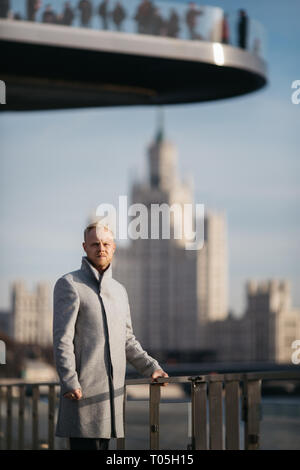 Bild des blonden Mann im grauen Mantel in der Stadt gegen den Hintergrund der Gebäude. Stockfoto