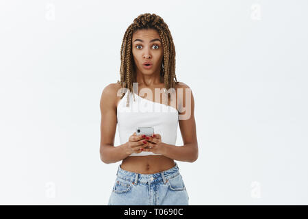 Innen- schuss von amüsiert enthusiastisch attraktive Afrikanische amerikanische Frau mit dreads in Weiß top Holding smartphone Falten Lippen in wow Sound stehend Stockfoto
