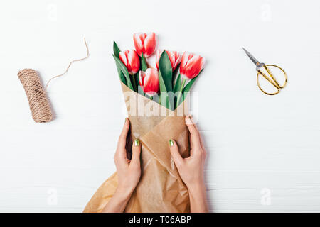 Der weibliche Hände wickeln Strauß roter Tulpen in Braun Kraftpapier neben Schere und Jute Seil auf weißem Holz- Tabelle, Ansicht von oben. Flach Zusammensetzung Stockfoto